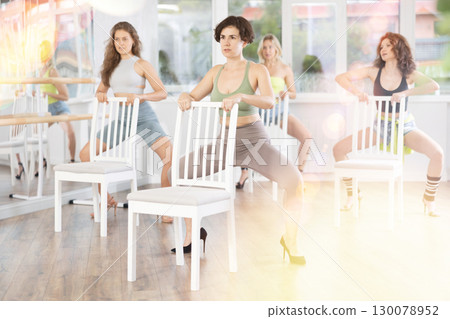 Cheerful young woman and multinational women training synchronous group choreography during dancing around chair and practicing strip plastic moves in dance studio 130078952