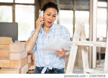 Asian woman with documents in her hands talking on mobile phone inside building under renovation 130078958