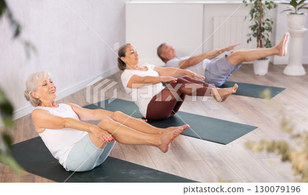 Class of elderly sporty women stretching gluteal muscle while lying on back during yoga Pilates workout on mats in gym studio 130079196