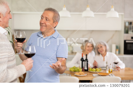 Elderly men have withdrawn from their female companions, stand with glass of wine in hands, chatting 130079201