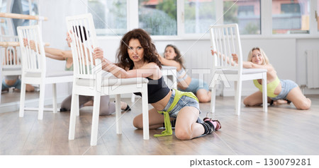 Young elegant female dancers in sportswear and high heels performing kneeling dance moves near chair on floor in choreographic studio during group lesson 130079281