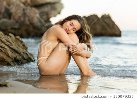 Semi naked young girl in bikini panties is sitting on sandy seashore 130079325