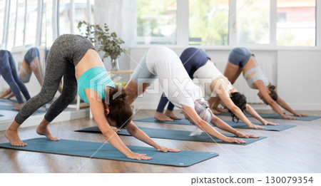 Woman performing stretching Adho Mukha Svanasana at group yoga class Woman performing stretching Adho Mukha Svanasana at group yoga class 130079354