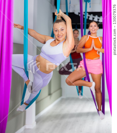 Young woman hanging upside down while practicing aerial yoga during group class in modern fitness club 130079376