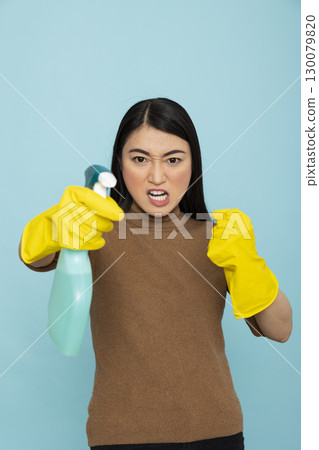Stressed asian housekeeper wearing rubber gloves and holding cleaning spray stands in aggressive fight pose against isolated background. Angry female cleaner showing frustration at house chores. 130079820