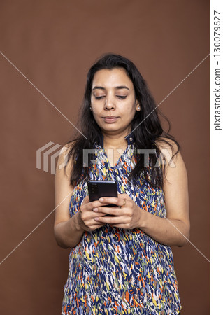 Indian woman using mobile device with focused expression, checking her emails and chats. Serious female individual standing and texting on smartphone against brown background studio. Indian woman using mobile device with focused expression, checking her emails and chats. Serious female individual standing and texting on smartphone against brown background studio. 130079827