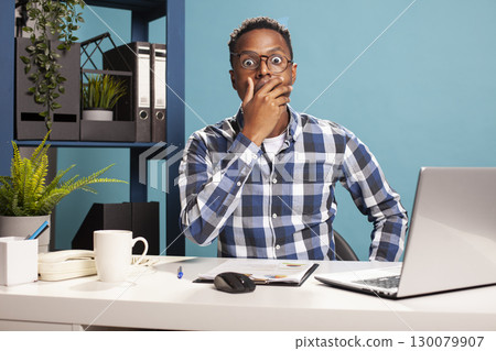 Company accountant covering his mouth in shock, surprised by misuse of business finances. Black employee stunned and perplexed by staff dismissal email, looking at camera with eyes wide open in fear. Company accountant covering his mouth in shock, surprised by misuse of business finances. Black employee stunned and perplexed by staff dismissal email, looking at camera with eyes wide open in fear. 130079907