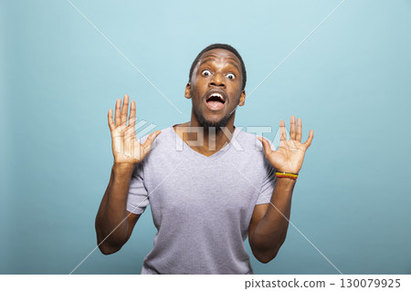 Astonished black male individual looking directly at the camera, raising both his hands in shock. Young african american man standing with a surprised expression on a blue background. Astonished black male individual looking directly at the camera, raising both his hands in shock. Young african american man standing with a surprised expression on a blue background. 130079925