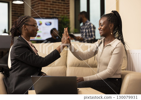 Two black businesswomen seated on couch, high fiving to celebrate success of a marketing plan. African american female employees rejoice after completing business project in startup office. 130079931