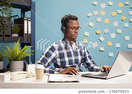 Financial consultant working on a laptop, analyzing business growth and sales management. Male secretary seated at desk with headset on, browsing on his device, preparing startup project presentation. Financial consultant working on a laptop, analyzing business growth and sales management. Male secretary seated at desk with headset on, browsing on his device, preparing startup project presentation. 130079933