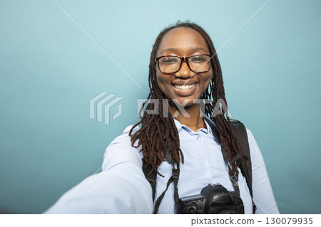 POV of trendy black woman grinning at camera while filming herself in a studio. African american female photographer stands with DSLR camera resting on her neck as she documents her creative process. POV of trendy black woman grinning at camera while filming herself in a studio. African american female photographer stands with DSLR camera resting on her neck as she documents her creative process. 130079935