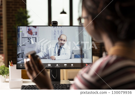 Female patient joins video call with doctor, watching medical consultation on desktop pc at home. Rear view of woman listening to male physician shown on computer screen during virtual appointment. 130079936