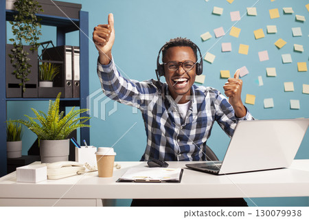 Smiling entrepreneur giving thumbs up and looking at camera, expressing happiness after successfully assisting a client. Cheerful man with headphones, excitedly showing approval gesture with his hands Smiling entrepreneur giving thumbs up and looking at camera, expressing happiness after successfully assisting a client. Cheerful man with headphones, excitedly showing approval gesture with his hands 130079938