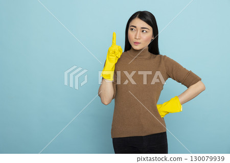 Young female housekeeper gestures upward with index finger, indicating isolated copy space for promotional use. Asian woman in rubber gloves poses against blue background, ready for cleaning job. Young female housekeeper gestures upward with index finger, indicating isolated copy space for promotional use. Asian woman in rubber gloves poses against blue background, ready for cleaning job. 130079939