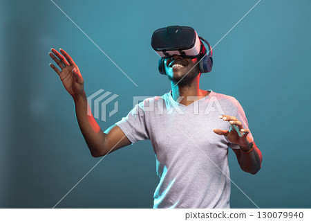Cheerful young man standing over blue background and experiencing an interactive 3D virtual simulation. Black male adult enjoying futuristic vision and tech, trying to touch something with his hands. Cheerful young man standing over blue background and experiencing an interactive 3D virtual simulation. Black male adult enjoying futuristic vision and tech, trying to touch something with his hands. 130079940