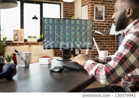 African american businessman seated at desk, analyzing financial charts on desktop pc. Male entrepreneur researching stock and crypto market trends to plan profitable investments for startup business. 130079942