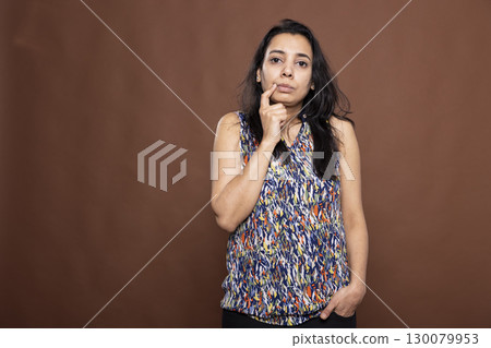 Gorgeous indian woman touching her chin, reflecting deeply while considering a response. Pensive female individual stands against brown background, conveying curiosity and uncertainty. Gorgeous indian woman touching her chin, reflecting deeply while considering a response. Pensive female individual stands against brown background, conveying curiosity and uncertainty. 130079953