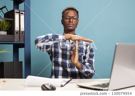 Male business consultant making a timeout symbol with his arms, signaling a break from work responsibilities. African american manager seated at office desk, using his hands to gesture a pause sign. Male business consultant making a timeout symbol with his arms, signaling a break from work responsibilities. African american manager seated at office desk, using his hands to gesture a pause sign. 130079954