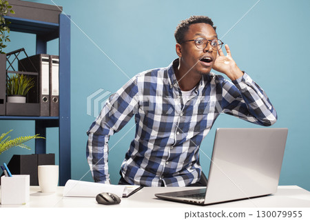 Male manager standing from his chair, mouth open and hand to his ear, trying to overhear something. Curious man is leaning forward from his table, wanting to hear clearly, interested in gossips. Male manager standing from his chair, mouth open and hand to his ear, trying to overhear something. Curious man is leaning forward from his table, wanting to hear clearly, interested in gossips. 130079955