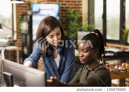 Caucasian manager attentively listening as black woman shares progress updates, gesturing to computer screen. Closeup of female coworkers collaborating on a startup project, discussing creative ideas. Caucasian manager attentively listening as black woman shares progress updates, gesturing to computer screen. Closeup of female coworkers collaborating on a startup project, discussing creative ideas. 130079990
