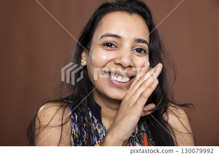 Closeup of attractive indian woman holding hand near mouth, playfully acting as if telling a secret or sharing gossip. Young lady smiling while whispering, isolated against brown background. Closeup of attractive indian woman holding hand near mouth, playfully acting as if telling a secret or sharing gossip. Young lady smiling while whispering, isolated against brown background. 130079992