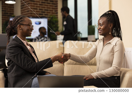 Cheerful female professionals sharing firm handshake, excited about completing financial report in brick wall office. African american businesswomen seated on couch, sealing a business project deal. 130080000