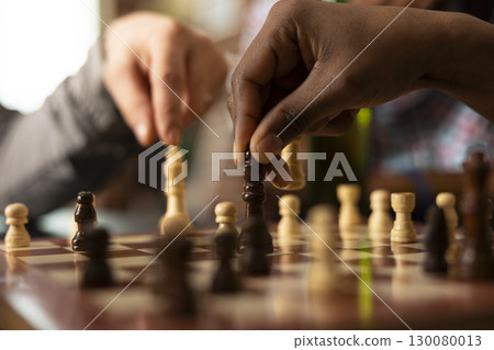 Selective focus of chess match in progress, capturing intensity of friendly game day. Close up of caucasian and african american individuals moving pieces on chessboard on table. 130080013