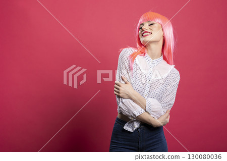 Portrait of cheerful caucasian woman with pink wig standing with eyes closed and arms crossed. Happy female individual posing against isolated background, expressing confidence and peace. Portrait of cheerful caucasian woman with pink wig standing with eyes closed and arms crossed. Happy female individual posing against isolated background, expressing confidence and peace. 130080036