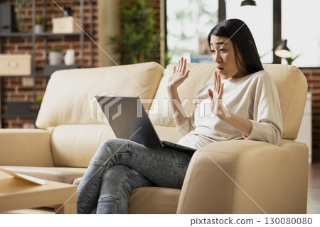 Remote female manager raises hands during online meeting, virtually presenting project ideas. Asian woman entrepreneur sits on sofa using laptop to pitch to potential investors from home. 130080080