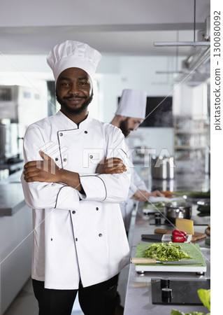 Portrait of african american head chef in a luxury restaurant kitchen works on food prep for cooking dishes, preparing fine dining meals for professional cuisine. Cook next to kitchen counter. 130080092