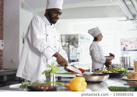 Black head chef chopping a fresh bell pepper in a modern workspace, preparing raw materials for a delicious gourmet recipe. Slicing veggies at the kitchen counter with utensils and cookware. 130080102