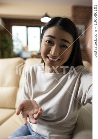 Young female adult recording business vlog on couch, sharing marketing strategies and investment tips. Smiling self employed woman speaking confidently to smartphone camera for social media audience. Young female adult recording business vlog on couch, sharing marketing strategies and investment tips. Smiling self employed woman speaking confidently to smartphone camera for social media audience. 130080103