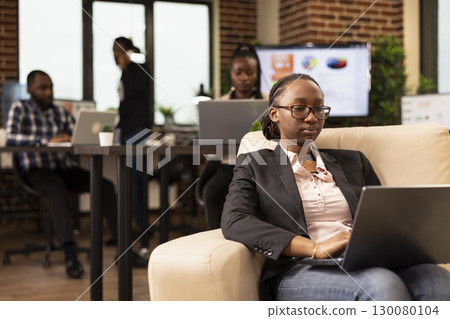 African american female entrepreneur working on laptop with focused expression, reviewing business analytics. Professional woman with glasses, siting on comfortable sofa and using personal computer. 130080104