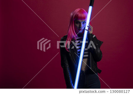 Young woman in striped crop top holds a glowing tube light her pink hair and leather jacket completing a moody, futuristic fashion shoot set in low light. Female model posing with led lamp stick. 130080127