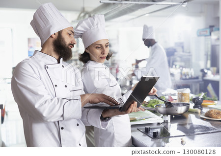 Team of two chefs researching recipes for new gourmet dishes on laptop, preparing meals in modern equipped restaurant kitchen. Professionals work on food prep, fresh produce ingredients. Team of two chefs researching recipes for new gourmet dishes on laptop, preparing meals in modern equipped restaurant kitchen. Professionals work on food prep, fresh produce ingredients. 130080128