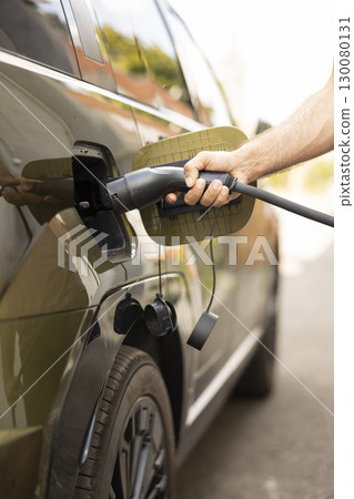 Close up of man connecting cable to EV vehicle inlet port charging at station. Driver opening charging port flap, plugging electric car into charger, reducing greenhouse gas emissions Close up of man connecting cable to EV vehicle inlet port charging at station. Driver opening charging port flap, plugging electric car into charger, reducing greenhouse gas emissions 130080131
