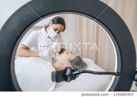 A cosmetologist blogger performs a procedure using a ring lamp to show subscribers the process of introducing fillers 130080407