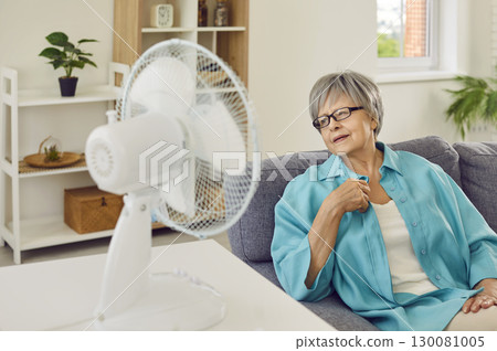 Senior woman suffering from summer heat and trying to cool down by electric fan at home 130081005