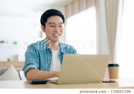 Happy young Asian man working on laptop in living room Happy young Asian man working on laptop in living room 130081596