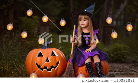 Little girl in witch costume sitting on pumpkin with broom during Halloween celebration with glowing lanterns. 130081851