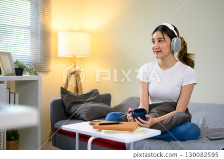 Relaxed young woman wearing headphones, sitting on sofa with coffee mug in a living room Relaxed young woman wearing headphones, sitting on sofa with coffee mug in a living room 130082595