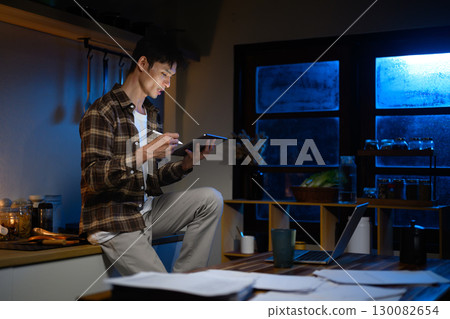 Overworked man working late at home using digital tablet sitting in the kitchen under warm lighting 130082654