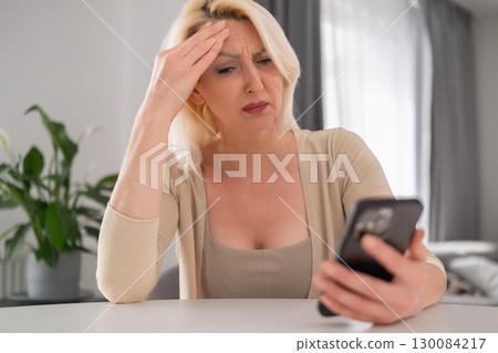 Blonde middle aged woman holding phone with tense grip, sitting in stylish apartment, looking at screen with distressed and anxious expression, symbolizing bad news or financial problem 130084217