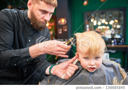 Professional hairdresser shaving boy's head, using shaver and comb. Cute boy excited about new haircut. Blond boy getting stylish haircut in barbershop. 130085845