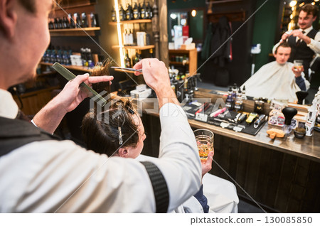 Back view of professional hairdresser styling hair. Professional stylist cutting hair while client enjoying his drink. Young customer sitting in front of mirror and getting new haircut. 130085850