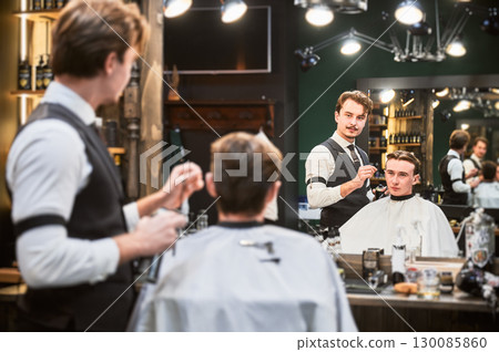 Professional barber showing result to client. Male customer looking at himself in mirror. Handsome hairdresser checking his reflection. 130085860