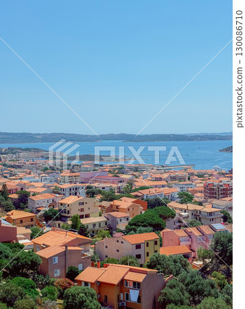 A stunning townscape showcasing charming buildings with red tiled roofs nestled by the coastline. The expansive view includes vivid blue waters and distant islands under a clear sky. Sardinia A stunning townscape showcasing charming buildings with red tiled roofs nestled by the coastline. The expansive view includes vivid blue waters and distant islands under a clear sky. Sardinia 130086710