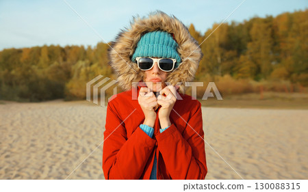 Woman is cold, freezing trying to warm up outdoors on beach on autumn day, wearing warm jacket 130088315