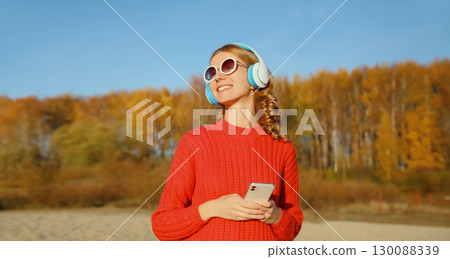 Happy young woman enjoying listening to music in headphones with phone outdoors in autumn park 130088339