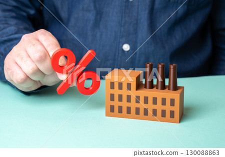 A man holds a percent sign near a factory. The industry is over-indebted. High interest rates are stifling business. Credit crisis, loan burden 130088863